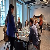 Auf diesem Bild ist eine Gruppe von Menschen zu sehen, die um einen Tisch sitzen, offenbar in einer Besprechung oder Diskussion. Auf dem Tisch liegen Papiere und Laptops, und der Raum hat große Fenster, durch die natürliches Licht einfällt. 