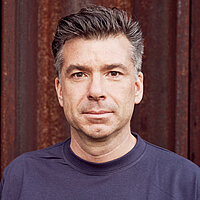 Ein Portraitbild. Ein Mann mit leicht gräulichen Haar blickt frontal in die Kamera. Er trägt einen blauen Pullover und steht vor einer braunen Wand.