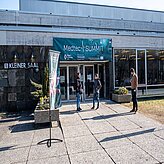 Das Bild zeigt den Eingang eines Gebäudes mit einem grünen Vordach, auf dem weißer Text in japanischer Sprache zu sehen ist. Vor dem Eingang stehen drei Personen. Links vom Eingang befindet sich ein Banner. Der Boden ist mit Steinen gepflastert, und vor dem Gebäude sind einige Pflanzen zu sehen. Das Gebäude selbst hat große Fenster und ein modernes Design.