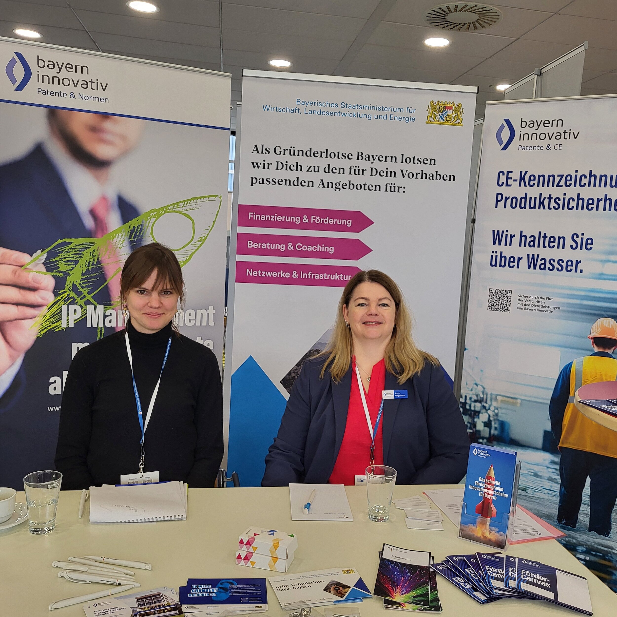 Charlotte Stegmayer, Projektmanagerin des Innovationsnetzwerk Kreativwirtschaft der Bayern Innovativ GmbH und Katrin Wägemann, Projektmanagerin Patente und CE der Bayern Innovativ GmbH, sitzen an einem Tisch. Vor ihnen liegen ausgebreitet etliche Flyer und Stifte. Im Hintergrund stehen drei Roll-ups.