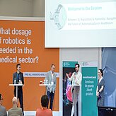 Eine Gruppe von Personen steht auf einer Bühne vor einem großen Bildschirm. Im Hintergrund sind Dekorationen und Banner zu sehen. 