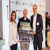 Drei Personen stehen nebeneinander und halten gemeinsam einen großen Scheck. Der Scheck trägt die Aufschrift '1st place' und '€ 12 000,-'. Im Hintergrund sind verschiedene Logos und Schriftzüge zu sehen.