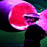 Eine Person trägt eine Virtual-Reality-Brille und blickt nach oben. Im Hintergrund sind große, leuchtende kugelförmige Installationen in Pink- und Violetttönen zu sehen.