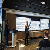 Präsentationsszene: Eine Person spricht vor einer Leinwand mit mehreren Folien, Teilnehmende hören zu. Das Foto zeigt einen Moment der Delegationsreise in Kanada.