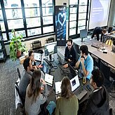 Das Bild zeigt junge Erwachsene, die an Laptops arbeiten und an einem Projekt im Bereich der digitalen Gesundheitsversorgung zusammenarbeiten. Im Hintergrund ist ein Poster mit dem Logo "digiCare" zu sehen. Die Atmosphäre ist konzentriert und kollaborativ, der Raum modern und hell.