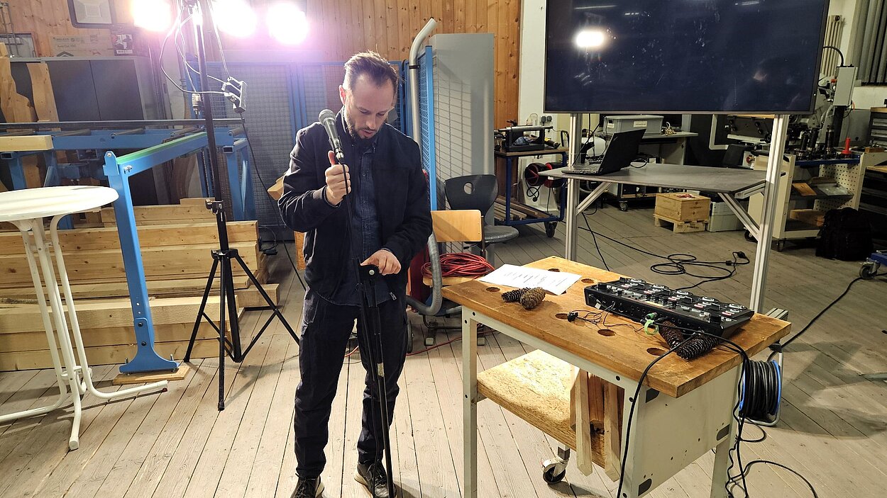 Ein Mann steht auf einer Art Bühne, mit Mikrofon in der Hand und neben ihm ein Mischpult. 