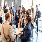 das Bild zeigt eine Gruppe von Personen bei einer gesellschaftlichen oder beruflichen Veranstaltung. Mehrere Personen stehen oder sitzen an Tischen und unterhalten sich. 