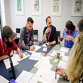 Mehrere Teilnehmende sitzen gemeinsam an einem Tisch und tauschen sich im Rahmen eines Workshops aus. Auf dem Tisch liegen Arbeitsunterlagen, Notizen und Schreibmaterialien.