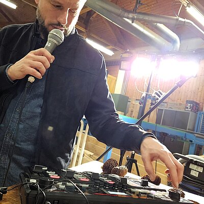 Ein Mann steht vor einen Mischpult und hält sich ein Mikrofon vor den Mund. 