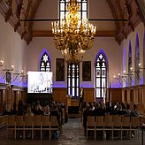 Ein großer Raum mit einem großen Kronleuchter und Menschen, die in Stühlen sitzen