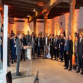 "Eine größere Gruppe von Personen befindet sich in einem geräumigen, gut beleuchteten Raum mit Holzsäulen und -balken. Die Anwesenden tragen formelle Kleidung. Eine Person steht an einem Rednerpult und spricht zum Publikum. Der Raum ist in warmem, orangefarbenem Licht ausgeleuchtet."