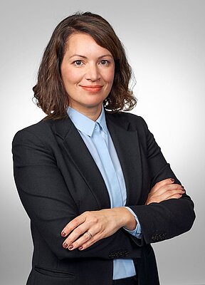 Person in Business-Outfit mit verschränkten Armen vor neutralem Hintergrund.