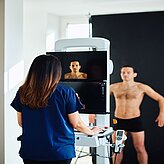 Person steht vor einem medizinischen Scanner, Fachkraft bedient das Gerät in einem hellen Raum.