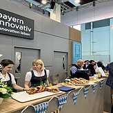 Auf dem Bild sind zwei Frauen zu sehen, welche hinter einer Theke stehen, welche in den bayerischen Farben geschmückt ist. Die Frauen tragen ein Dirndl und geben Speisen aus.