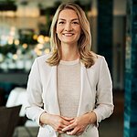 Eine Person in einem hellen Blazer über einem gemusterten Oberteil steht in einem Innenraum, der möglicherweise ein Restaurant oder Café darstellt.