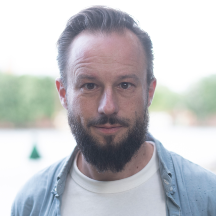 Ein Mann mit weißen T-Shirt, Jeanshemd und Bart schaut frontal in die Kamera.