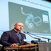 das Bild zeigt eine Person an einem Rednerpult, die eine Präsentation hält.