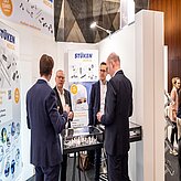 "Vier Männer in Businesskleidung stehen an einem Messestand und unterhalten sich. Der Stand hat einen weißen Hintergrund mit dem Firmennamen 'LUMEN' sowie Produktabbildungen und Beschreibungen. Auf dem Tisch vor ihnen befinden sich verschiedene Gegenstände, vermutlich im Zusammenhang mit den ausgestellten Produkten."