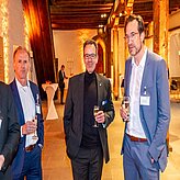 Das Bild zeigt drei Personen in formeller Kleidung, die bei einer gesellschaftlichen oder Netzwerkveranstaltung stehen und Getränke in der Hand halten. Der Hintergrund ist von warmem Licht erhellt und zeigt einen Raum mit hoher Decke und Holzbalken.