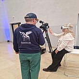 In einem Innenraum sitzt eine Frau mit einer VR-Brille und schaut in eine Kamera, während ein Mann neben ihr steht.