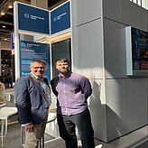 Auf dem Bild sind zwei Männer zu sehen, welche vor einer Werbewand stehen. 