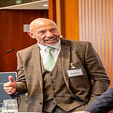 Das Bild zeigt eine Person in einem braunen, gemusterten Anzug mit weißem Hemd und hellgrüner Krawatte. Die Person trägt ein Namensschild am Jackett und hält eine Hand mit ausgestrecktem Daumen nach oben. 