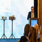 Tobias Gotthardt, Staatssekretär im Bayerischen Staatsministerium für Wirtschaft, Landesentwicklung und Energie, steht an einem Rednerpult und spricht vor Publikum. Neben ihm befinden sich zwei Stehtische mit goldfarbenen Trophäen.