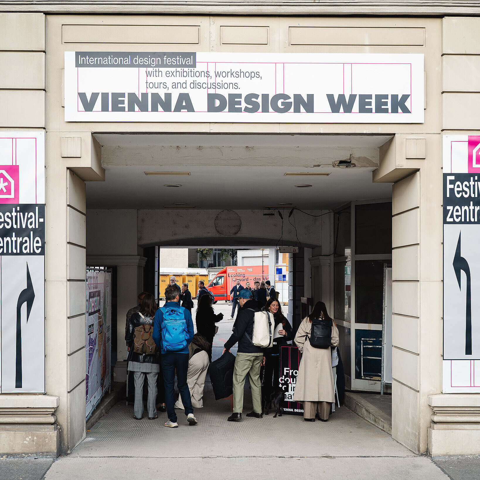 Mehrere Menschen stehen in einem überdachten Durchgang unter einem Schild mit der Aufschrift „Vienna Design Week“. Der Durchgang führt zu einer Straße, wo man im Hintergrund weitere Passanten sieht.