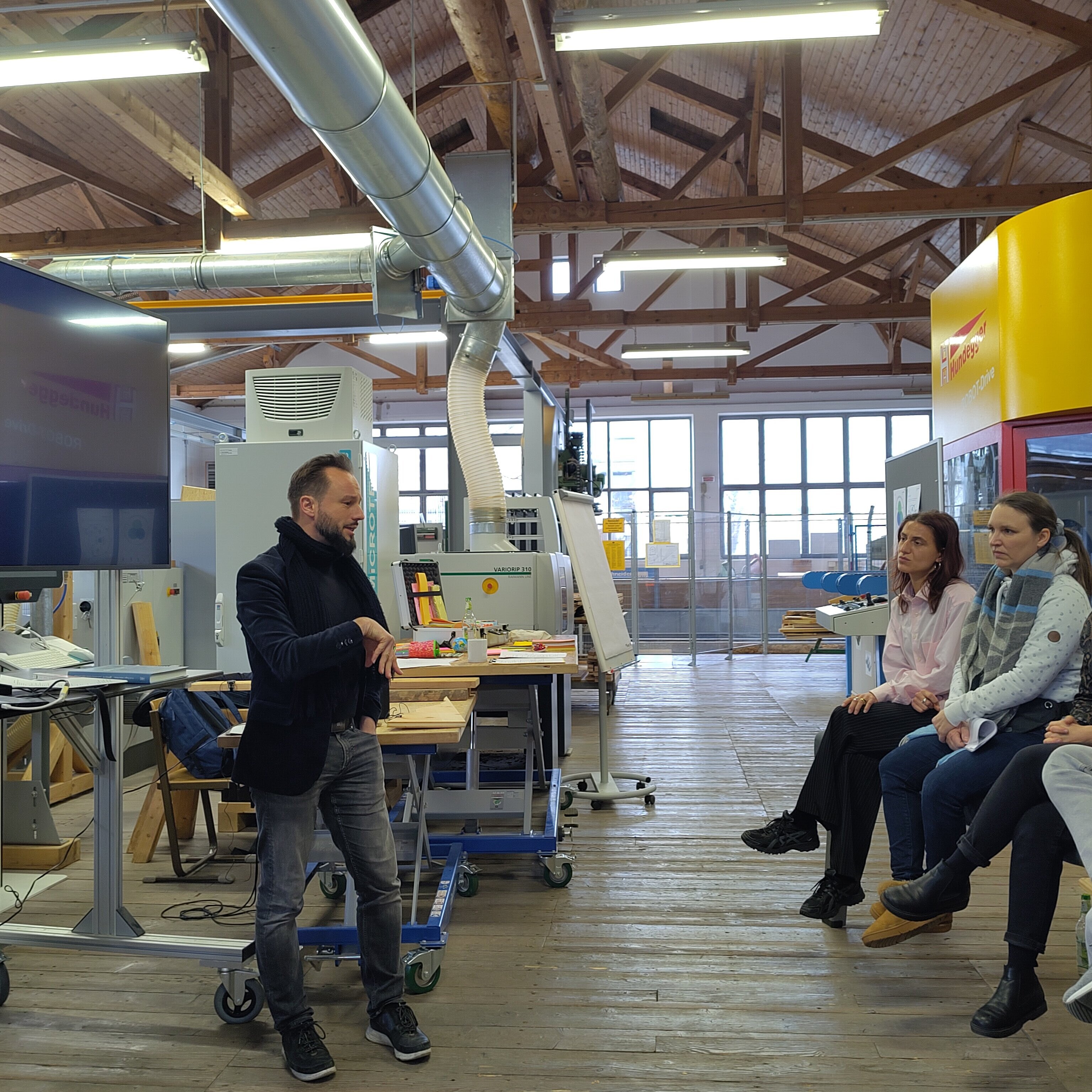 Blick in ein Sägewerk. Ein Mann links im Bild erklärt etwas vor einem kleinen Publikum.