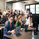 Ergebnis der Bildbeschreibung Das Bild zeigt eine Gruppe von Menschen, die in einem hellen Raum an Laptops sitzen. Es scheint sich um eine Art Workshop oder Konferenz zu handeln, da die Teilnehmer konzentriert an ihren Geräten arbeiten und viele Laptops und Computer auf den Tischen stehen. Im Hintergrund sind weitere Menschen zu sehen, die ebenfalls an der Veranstaltung teilnehmen. Die Atmosphäre wirkt konzentriert und produktiv. Die meisten Personen tragen eher legere Kleidung.