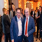 "Mehrere Personen befinden sich bei einer formellen Veranstaltung in einem Raum mit warmem, orangefarbenem Licht. Zwei Männer im Vordergrund posieren für das Foto, beide tragen Anzug und Krawatte. Weitere Personen sind im Hintergrund zu sehen."