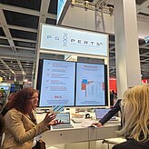 Auf dem Bild sind zwei Frauen zu sehen, welche in einem Gespräch verwickelt sind. Im Hintergrund ist ein Messestand mit Bildschirmen zu sehen.