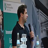 Eine Person spricht bei einer Veranstaltung in ein Mikrofon. Im Hintergrund ist ein grün-weißer Banner mit teilweise unleserlichem Text zu sehen. Die Person sitzt an einem Tisch, auf dem mehrere Wasserflaschen stehen.