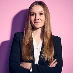 Auf dem Bild ist eine Frau mit langen Haaren zu sehen. Sie trägt einen schwarzen Hosenanzug.