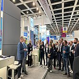 Auf dem Bild ist eine Gruppe von Personen in einem Veranstaltungsraum zu sehen, welche sich stehend einen Vortrag anhören.