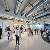 Das Bild zeigt eine Veranstaltung in einem modernen, hellen Raum mit hohen Decken und sichtbarer Industrie-Rohrleitung. Mehrere Personen stehen und sitzen im Raum, manche hören einem Redner zu, der an einem Stehpult steht. Im Hintergrund steht ein Banner von "3D Systems", welches auf eine Partnerschaft am Point of Care hinweist.