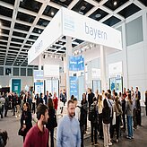 Messestand mit der Aufschrift »bayern« auf einem großen weißen Banner über einem belebten Ausstellungsbereich mit vielen Menschen in einer Halle