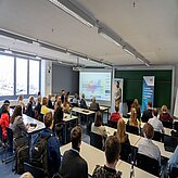 Vortragender steht vor Projektionsleinwand in einem vollbesetzten Seminarraum mit Zuhörern an Tischen