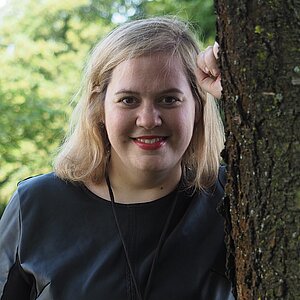 Eine Frau mit blonden, schulterlangen Haaren steht von hinten an einen Baum angelehnt, während sie freundlich und aufgeschlossen in die Kamera lächelt. Sie trägt dunkelblaue Kleidung, roten Lippenstift und eine Kette mit einem markanten goldenen Anhänger. 
