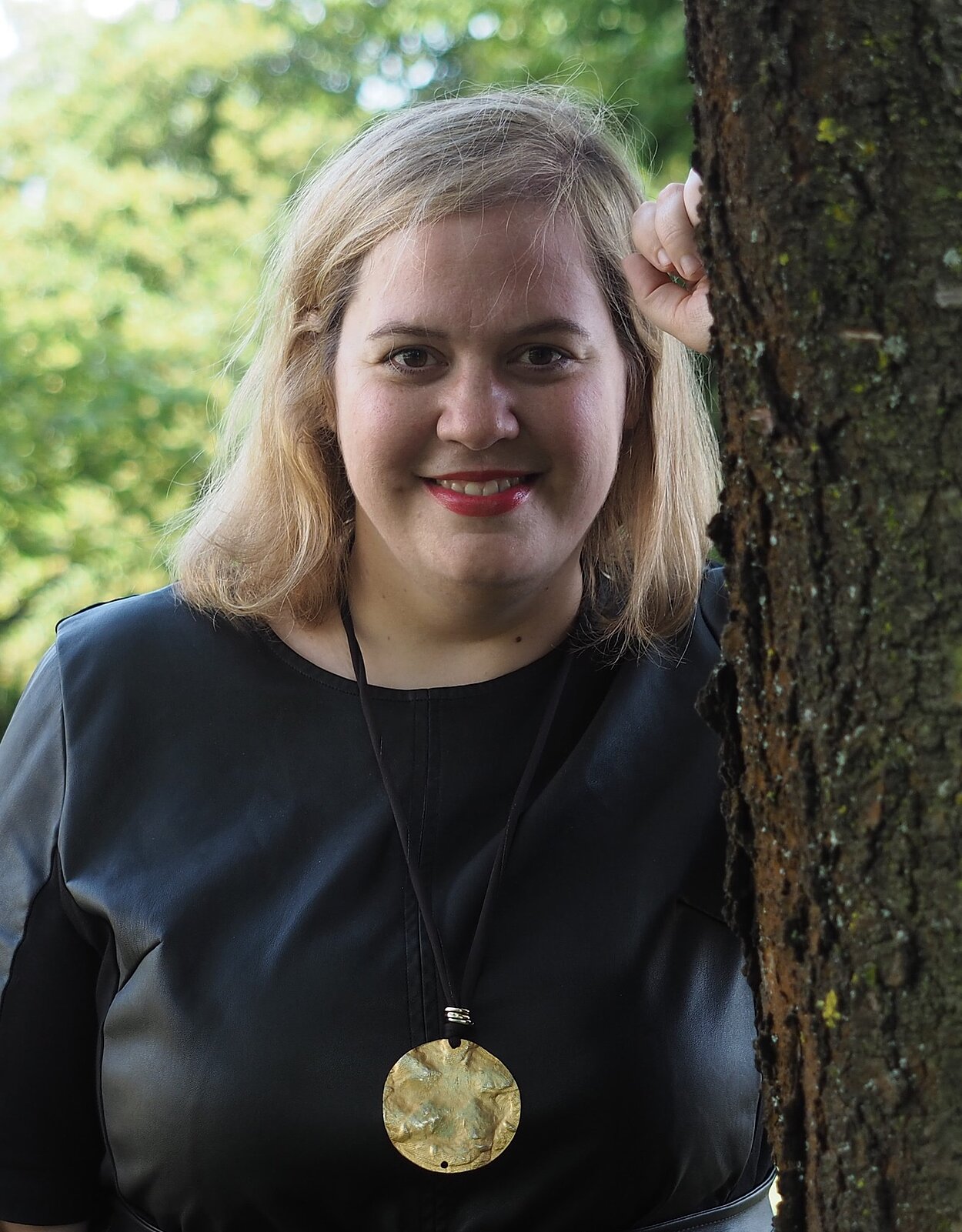 Eine Frau mit blonden, schulterlangen Haaren steht von hinten an einen Baum angelehnt, während sie freundlich und aufgeschlossen in die Kamera lächelt. Sie trägt dunkelblaue Kleidung, roten Lippenstift und eine Kette mit einem markanten goldenen Anhänger. 