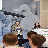 Bei einer öffentlichen Veranstaltung oder Präsentation stehen zwei Personen im Vordergrund, eine spricht in ein Mikrofon, die andere sitzt hinter einem Rednerpult