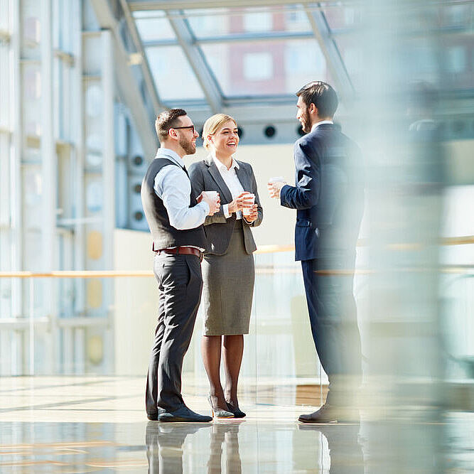 Zwei Männer und eine Frau im Business Look, unterhalten sich in einer verglasten Halle
