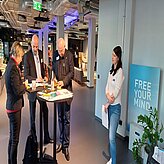 Auf diesem Bild ist eine Gruppe von Menschen zu sehen, die um hohe Tische stehen, möglicherweise bei einer sozialen oder Networking-Veranstaltung. Eine weitere Person steht in der Nähe einer Wand, hinter der sich ein großes Poster oder eine Anzeige befindet.