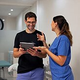 Eine Frau in blauer medizinischer Arbeitskleidung spricht mit einem Mann in schwarzem T-Shirt, der ein Tablet hält, in einem hellen Raum mit medizinischer Ausstattung.