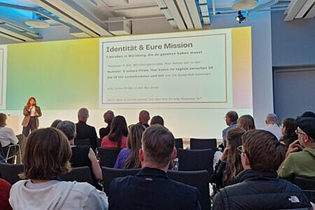 Auf der Bühne der Transformaing Media ist Caroline Mohr zu sehen, wie sie gerade einen Vortrag hält. Der bestuhlte Saal ist gut besucht und es sind alle Plätze besetzt. Von den Zuhörenden sind nur die Hinterköpfe zu sehen, während die Referentin selbst nur dezent aus der Ferne zu sehen ist.