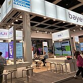 Auf dem Bild ist ein Messestand mit weißen Messemöbeln und verschiedenen Aufstellern zu sehen.