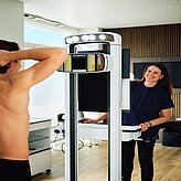 Medizinische Untersuchungsszene mit einer stehenden Person in Unterwäsche vor einem schwarzen Hintergrund und einer Fachkraft an einem Gerät.