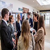 Gruppe von Personen bei einer gesellschaftlichen oder beruflichen Veranstaltung. Mehrere Personen stehen oder sitzen an Tischen und unterhalten sich. 