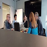 Auf diesem Bild sind fünf Personen zu sehen, die um einen Tisch sitzen, offenbar in einer Besprechung oder Diskussion. Auf dem Tisch stehen einige Flaschen und Gläser, und an der Wand hinter ihnen ist ein großer Bildschirm angebracht.
