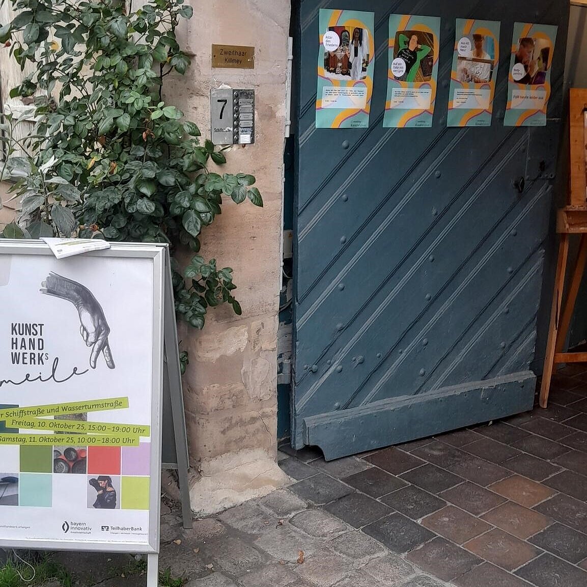 Geöffnete türkise Tür mit Postern, davor steht ein Posteraufsteller. 